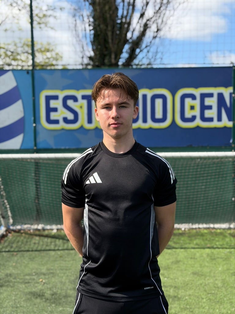 Young male athlete wearing black Adidas shirt standing on tennis court with blue barrier and trees in background