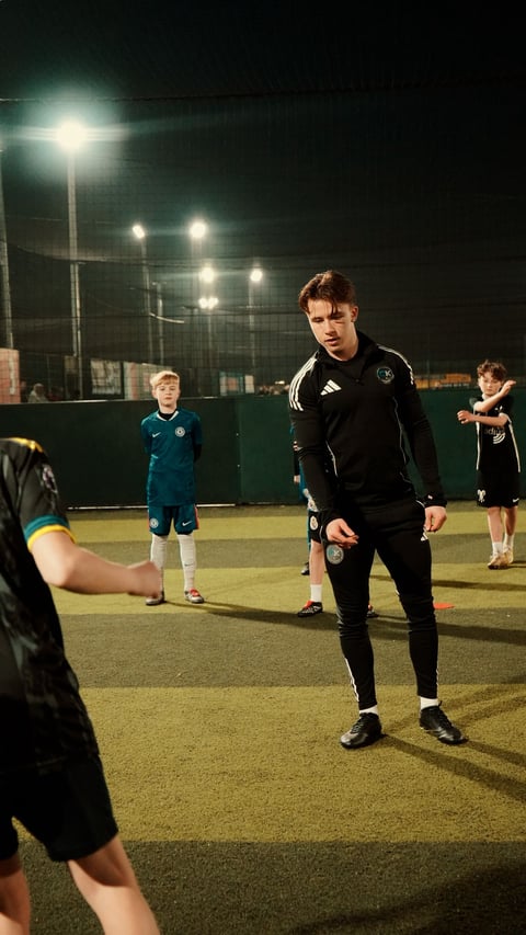 Young soccer coach in black Adidas jacket demonstrating skills to children on a lit nighttime artificial grass field